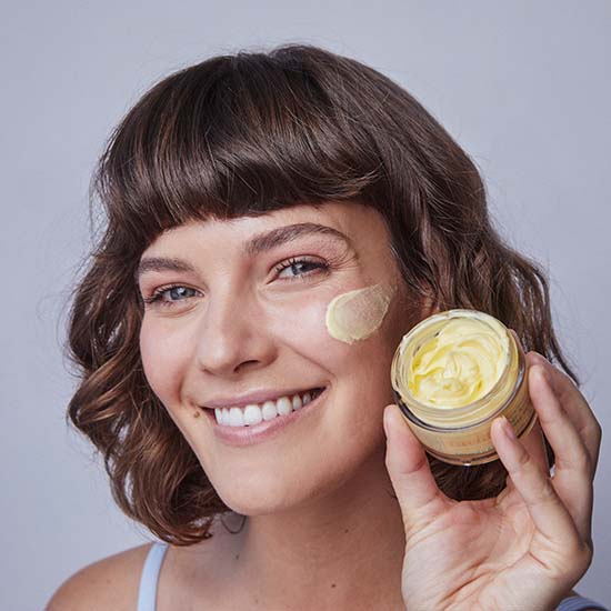 Kobieta z kremem Age Away Hydrating Cream Lavido nałożonym na policzek, trzyma otwarty słoiczek